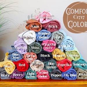 May include: A pyramid of rolled t-shirts in various colors, including white, red, blue, green, yellow, and brown. The t-shirts are labeled with color names, such as "White", "Red", "Blue", "Green", "Yellow", and "Brown". The text "COMFORT C1717 COLORS" is displayed in a circle at the top of the image.
