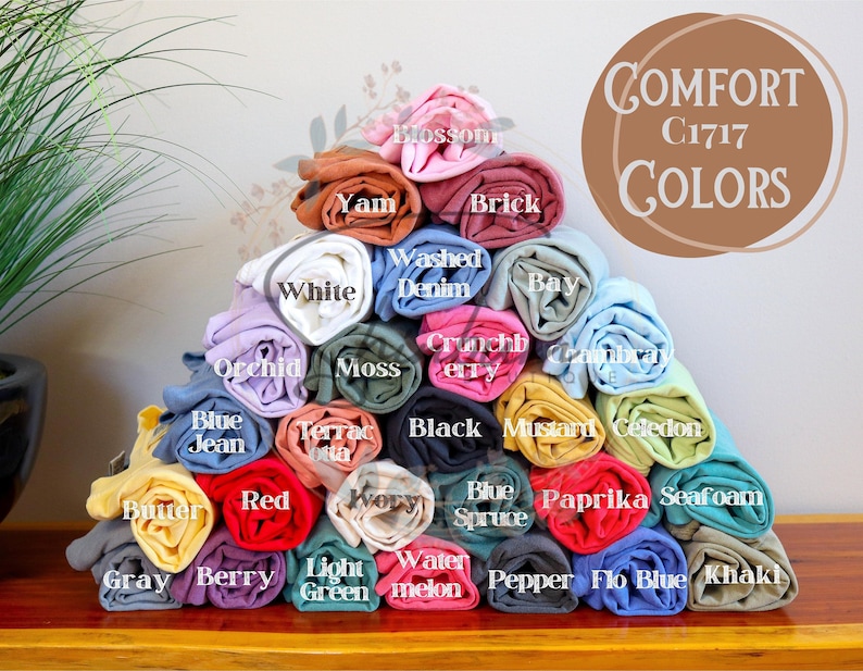 May include: A pyramid of rolled t-shirts in various colors, including white, red, blue, green, yellow, and brown. The t-shirts are labeled with color names, such as "White", "Red", "Blue", "Green", "Yellow", and "Brown". The text "COMFORT C1717 COLORS" is displayed in a circle at the top of the image.