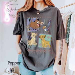 May include: Dark gray t-shirt featuring a colorful graphic of characters from The Lion King. The design includes Simba, Timon, Pumbaa, and Zazu, set against a patterned background. The word "Pepper" is visible on the shirt.