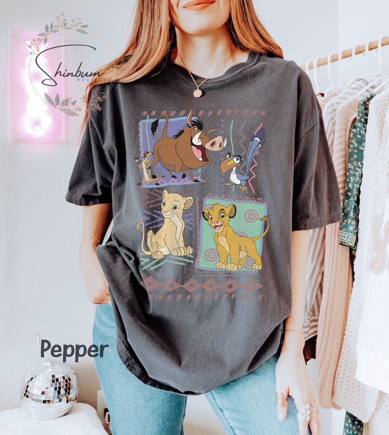 May include: Dark gray t-shirt featuring a colorful graphic of characters from The Lion King. The design includes Simba, Timon, Pumbaa, and Zazu, set against a patterned background. The word "Pepper" is visible on the shirt.