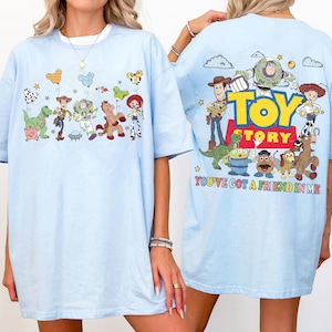 May include: A light blue t-shirt featuring Toy Story characters. The front displays Rex, Woody, Buzz Lightyear, Bullseye, and Jessie with Mickey-shaped balloons. The back shows the Toy Story logo and characters with the text You've Got A Friend In Me.