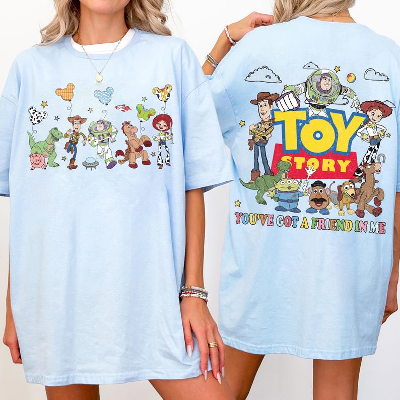 May include: A light blue t-shirt featuring Toy Story characters. The front displays Rex, Woody, Buzz Lightyear, Bullseye, and Jessie with Mickey-shaped balloons. The back shows the Toy Story logo and characters with the text You've Got A Friend In Me.