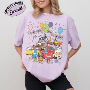 K&ouml;nnte beinhalten: Ein lila T-Shirt mit einem farbenfrohen Design von Cartoon-Autos, Luftballons und Geburtstagsdekorationen. Das Design enth&auml;lt den Text "HAPPY BIRTHDAY" und Platz f&uuml;r einen Namen. Das Shirt zeigt auch den Text "COMFORT COLORS" und die Farbe "Orchid".