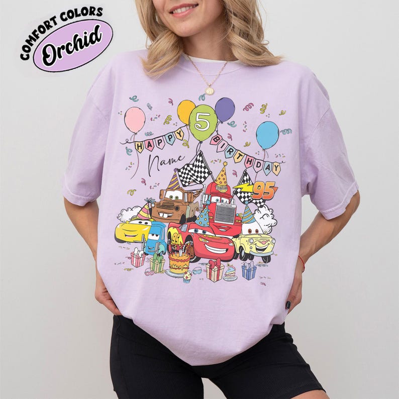K&ouml;nnte beinhalten: Ein lila T-Shirt mit einem farbenfrohen Design von Cartoon-Autos, Luftballons und Geburtstagsdekorationen. Das Design enth&auml;lt den Text "HAPPY BIRTHDAY" und Platz f&uuml;r einen Namen. Das Shirt zeigt auch den Text "COMFORT COLORS" und die Farbe "Orchid".