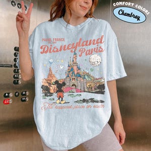 May include: Light blue t-shirt featuring a graphic of Disneyland Paris with Mickey Mouse. The shirt has the text "Paris, France" and "Disneyland Paris" in a vintage style. The phrase "The happiest place on earth" is also printed on the shirt.