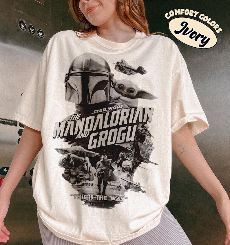 Op de afbeelding: Ivoor kleurig T-shirt met een afbeelding van personages uit de serie "The Mandalorian", waaronder de Mandalorian en Grogu. Het ontwerp bevat de tekst "The Mandalorian and Grogu" en "This is the Way."