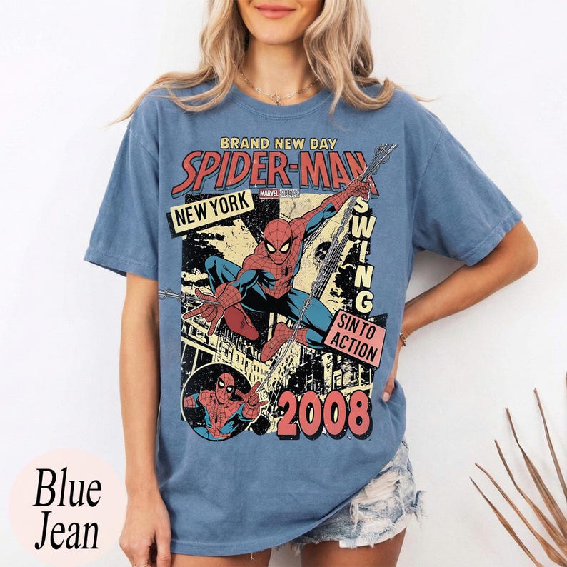 May include: Light blue t-shirt with a vintage Spider-Man graphic. The design includes the words "Brand New Day," "Spider-Man," "New York," and "2008." The graphic shows Spider-Man swinging. The shirt is a light blue colour.