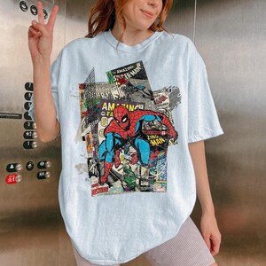 Pode incluir: Camiseta azul clara com um design de colagem de quadrinhos com o Homem-Aranha. O design inclui as palavras "AMAZING SPIDER-MAN" e "ZING MAN" em estilo de quadrinhos. A camiseta tem um caimento folgado e grande.