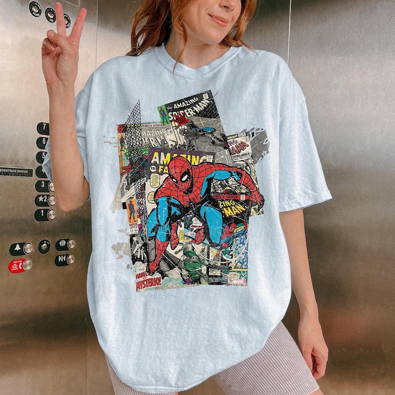 Pode incluir: Camiseta azul clara com um design de colagem de quadrinhos com o Homem-Aranha. O design inclui as palavras "AMAZING SPIDER-MAN" e "ZING MAN" em estilo de quadrinhos. A camiseta tem um caimento folgado e grande.