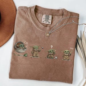 Puede incluir: Camiseta Comfort Colors marr&oacute;n con un gr&aacute;fico de Baby Yoda en varias poses. La camiseta tiene cuello redondo y mangas cortas. Un collar plateado con un colgante est&aacute; sobre la camiseta.