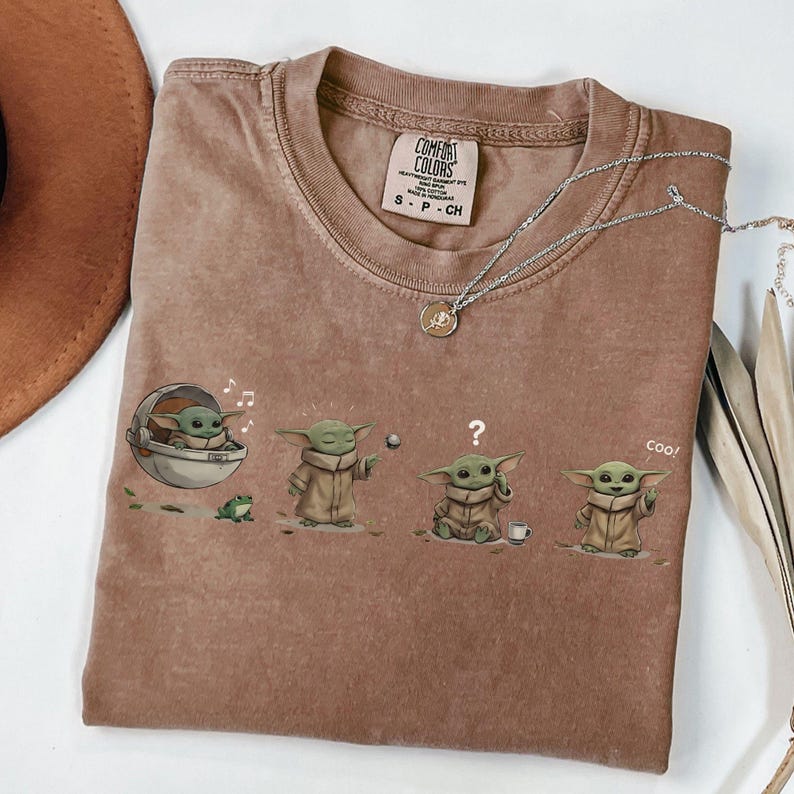 Puede incluir: Camiseta Comfort Colors marr&oacute;n con un gr&aacute;fico de Baby Yoda en varias poses. La camiseta tiene cuello redondo y mangas cortas. Un collar plateado con un colgante est&aacute; sobre la camiseta.