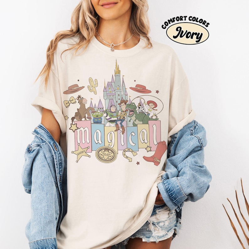 Puede incluir: Camiseta de color marfil con un gr&aacute;fico colorido que presenta personajes de dibujos animados, un castillo y la palabra "m&aacute;gico". La camiseta tiene un corte holgado y mangas cortas. El texto "Comfort Colors" e "Ivory" est&aacute; impreso en la parte superior.