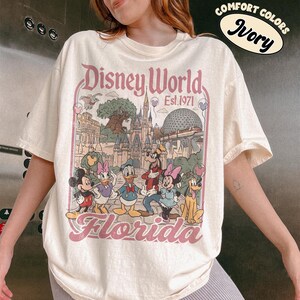May include: Ivory-colored Disney World Florida t-shirt featuring a vintage-style graphic of Disney characters and park landmarks. The design includes Mickey Mouse, Donald Duck, and Cinderella's castle. The text "Disney World Est. 1971" is also visible.