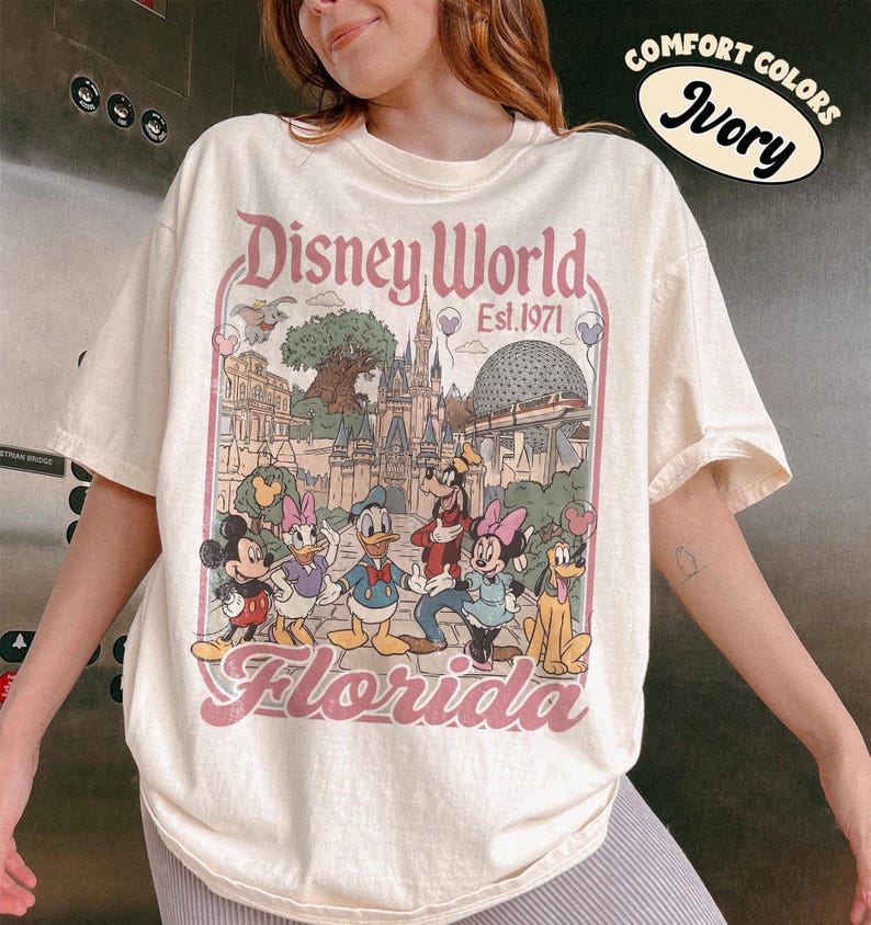 May include: Ivory-colored Disney World Florida t-shirt featuring a vintage-style graphic of Disney characters and park landmarks. The design includes Mickey Mouse, Donald Duck, and Cinderella's castle. The text "Disney World Est. 1971" is also visible.
