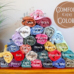 May include: A pyramid of rolled-up t-shirts in various colors, including white, blue, red, and green. The image displays the Comfort C1717 Colors, with each shirt labeled with its color name. The shirts are stacked on a wooden surface.