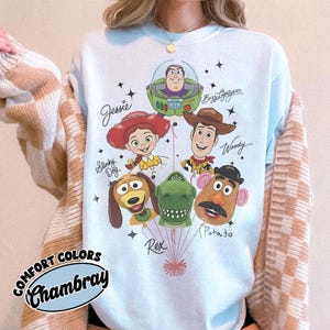 Puede incluir: Sudadera azul claro con personajes de dibujos animados, incluyendo a Jessie, Buzz Lightyear, Woody, Slinky Dog, Rex y Mr. Potato Head. El texto "Comfort Colors Chambray" est&aacute; en un dise&ntilde;o circular.