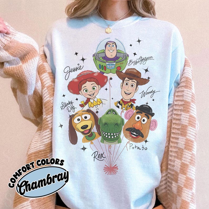 Puede incluir: Sudadera azul claro con personajes de dibujos animados, incluyendo a Jessie, Buzz Lightyear, Woody, Slinky Dog, Rex y Mr. Potato Head. El texto "Comfort Colors Chambray" est&aacute; en un dise&ntilde;o circular.