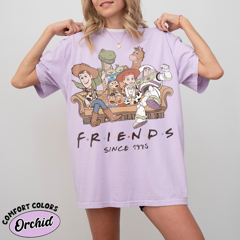 May include: A light purple short-sleeve t-shirt featuring a graphic print of various animated characters sitting on a couch. Below the characters, the text reads FRIENDS SINCE 1995. The shirt has a crew neckline and a relaxed, loose-fitting silhouette.