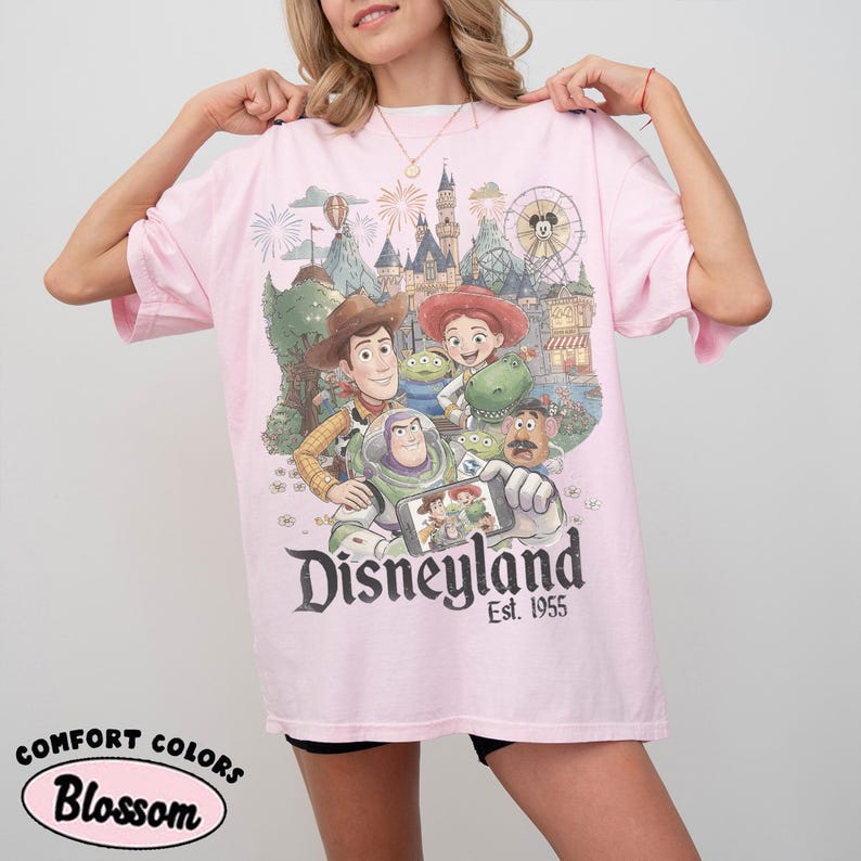 May include: A light pink short-sleeve t-shirt featuring a graphic print of Toy Story characters including Woody, Buzz Lightyear, and Jessie in front of a Disneyland castle. The text Disneyland Est. 1955 is printed below the colorful character illustration.