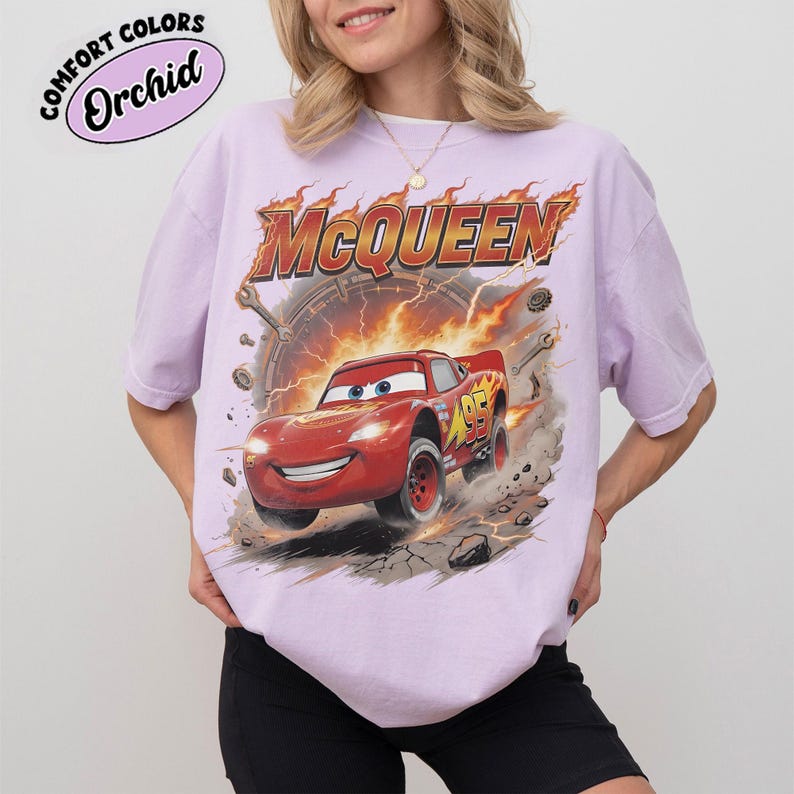 Może przedstawiać: Jasnofioletowa koszulka z grafiką Zygzaka McQueena z filmu Auta. Projekt zawiera napis "McQueen" w kolorze pomarańczowym i czerwonym, płomienne tło oraz numer 95 na samochodzie.