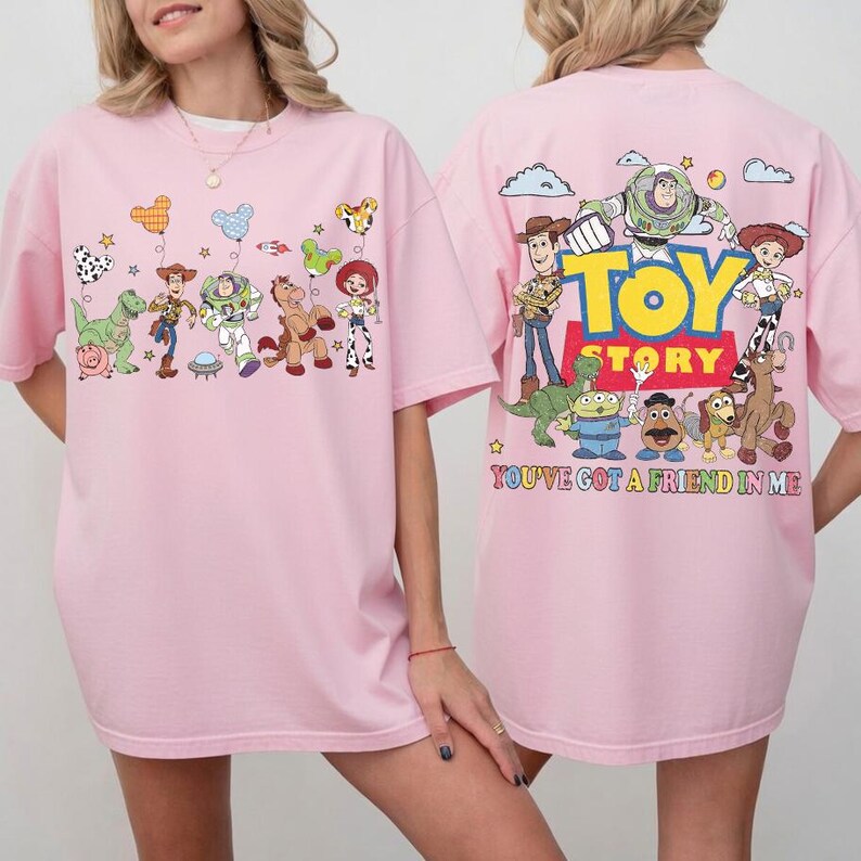 May include: A light pink t-shirt featuring Toy Story characters. The front displays various characters with balloons. The back shows the Toy Story logo, characters, and the text "YOU'VE GOT A FRIEND IN ME" in colorful letters on a light pink background.