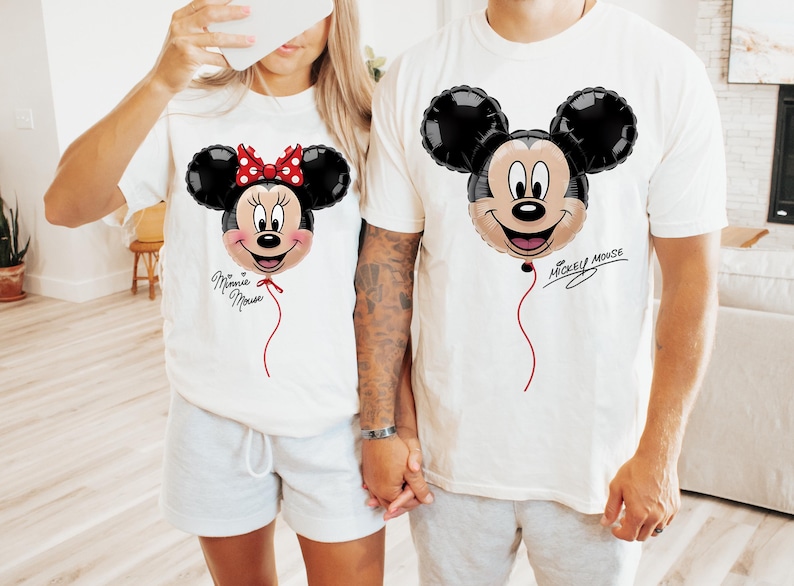 K&ouml;nnte beinhalten: Wei&szlig;e T-Shirts mit Mickey- und Minnie-Mouse-Ballon-Designs. Das Minnie-Mouse-Shirt hat eine rote Schleife und den Text "Minnie Mouse". Das Mickey-Mouse-Shirt hat den Text "Mickey Mouse". Beide Shirts haben eine rote Schnur am Ballon.