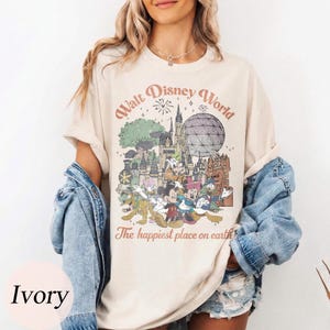 May include: Ivory-colored t-shirt featuring a vintage-style graphic of Walt Disney World. The design includes iconic landmarks, characters, and the text "Walt Disney World" and "The happiest place on earth" in a retro font.