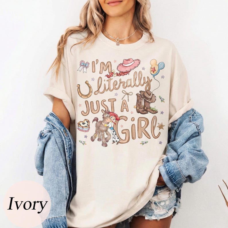 K&ouml;nnte beinhalten: Elfenbeinfarbenes T-Shirt mit dem Text "I'm Literally Just a Girl" und Cartoon-Grafiken eines Hufeisens, Cowboy-Stiefeln, einem Pferd und einem rosa Cowboyhut. Das Shirt hat eine lockere Passform.