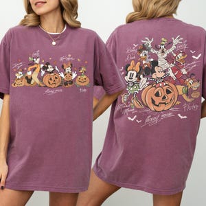May include: A mauve-colored t-shirt featuring a Halloween-themed graphic of Disney characters, including Mickey and Minnie Mouse, Goofy, Donald Duck, and Pluto, with carved pumpkins. The back of the shirt also displays the same characters with pumpkins. The text "Mickey Mouse" is visible.