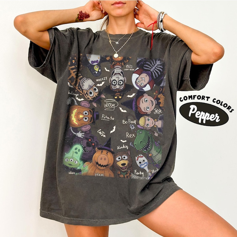 May include: A charcoal gray short-sleeve t-shirt featuring a graphic print of various animated characters dressed in Halloween costumes. The design includes text labels for each character and the words Comfort Colors Pepper on the side.