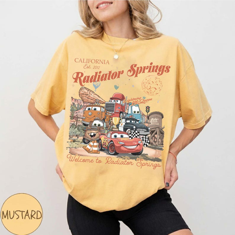 以下が含まれることがあります： マスタードイエローのTシャツ。映画「カーズ」のキャラクターのグラフィックが特徴で、「California Est. 2012 Radiator Springs」の文字が入っています。デザインには、さまざまな車のキャラクターと「Welcome to Radiator Springs」というフレーズが含まれています。