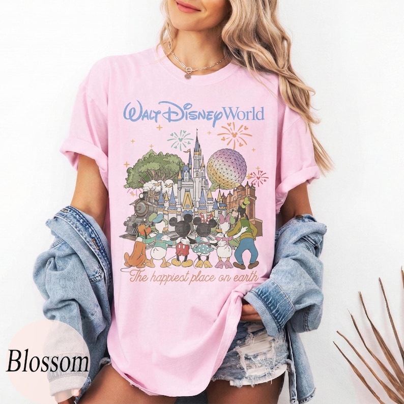 Puede incluir: Camiseta rosa claro con un gr&aacute;fico colorido de personajes y lugares emblem&aacute;ticos de Disney, incluyendo un castillo, un tren y Spaceship Earth. El texto "Walt Disney World" y "The happiest place on earth" est&aacute; impreso en la camiseta.