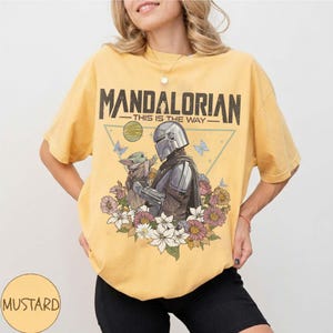 May include: Mustard yellow t-shirt featuring a graphic of The Mandalorian and Grogu surrounded by flowers and butterflies. The text "MANDALORIAN - THIS IS THE WAY" is printed across the top of the design. The shirt is a relaxed fit.