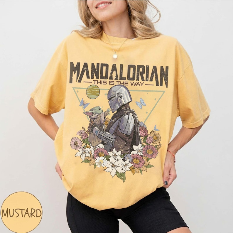May include: Mustard yellow t-shirt featuring a graphic of The Mandalorian and Grogu surrounded by flowers and butterflies. The text "MANDALORIAN - THIS IS THE WAY" is printed across the top of the design. The shirt is a relaxed fit.