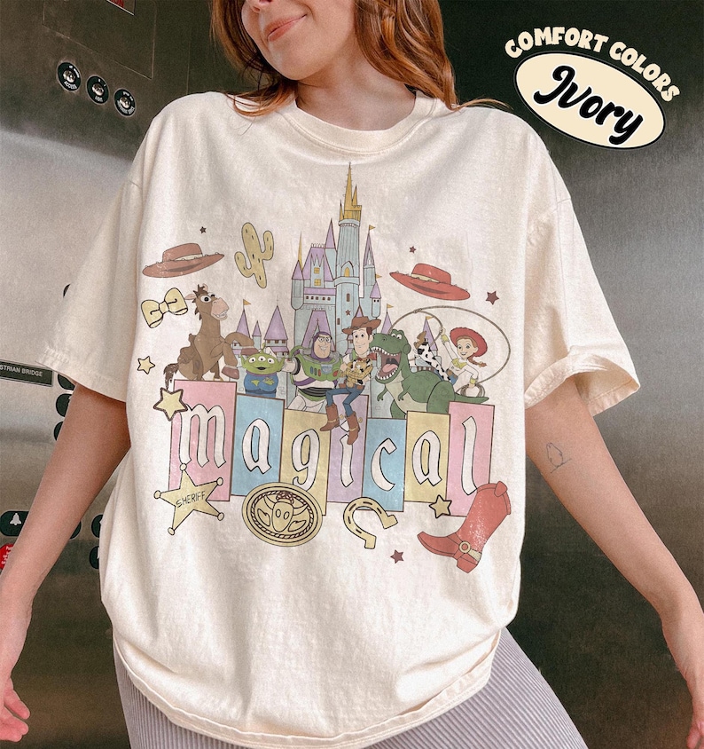 May include: Ivory-colored t-shirt featuring a colorful graphic of characters and elements from a popular animated movie. The word "magical" is displayed in large, colorful letters. The shirt also has the words "Comfort Colors" and "Ivory" printed on it.