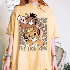May include: Mustard-colored t-shirt featuring a graphic of Simba, Pumbaa, and Timon from The Lion King, with the text "THE LION KING". The design is set against a brown and tan patterned background.
