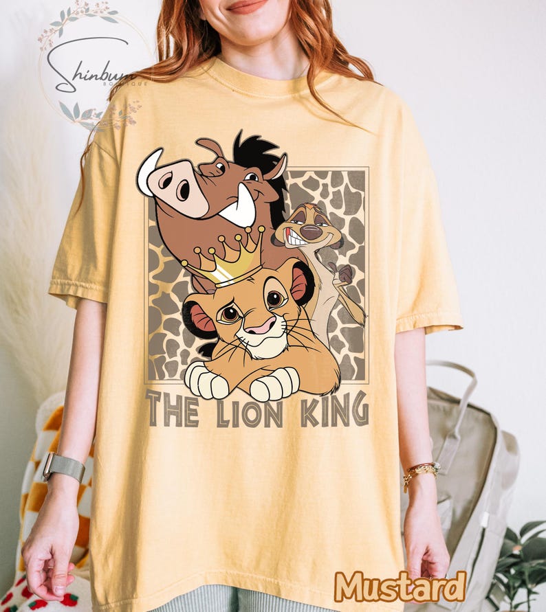 May include: Mustard-colored t-shirt featuring a graphic of Simba, Pumbaa, and Timon from The Lion King, with the text "THE LION KING". The design is set against a brown and tan patterned background.