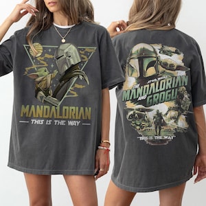 May include: Dark gray t-shirt featuring a graphic of The Mandalorian and Grogu. The front design includes the text "MANDALORIAN - THIS IS THE WAY" and a triangular graphic. The back design features the text "STAR WARS THE MANDALORIAN AND GROGU - THIS IS THE WAY".