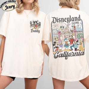 May include: Ivory-colored t-shirt featuring a Mickey Mouse graphic with the word "Daddy" on the front. The back of the shirt displays a Disneyland California design with various Disney characters and the text "Est. 1955".