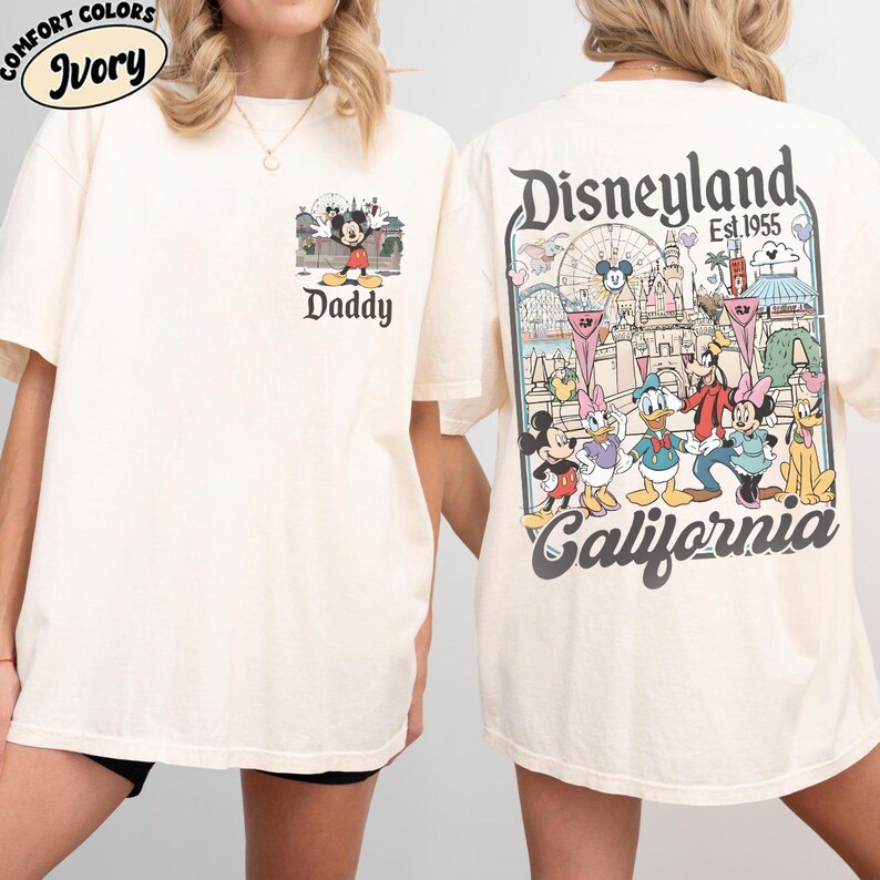 May include: Ivory-colored t-shirt featuring a Mickey Mouse graphic with the word "Daddy" on the front. The back of the shirt displays a Disneyland California design with various Disney characters and the text "Est. 1955".