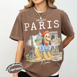 K&ouml;nnte beinhalten: Braunes T-Shirt mit dem Wort "PARIS" in wei&szlig;en Buchstaben und einer Grafik von Pluto vor Pariser Wahrzeichen. Das Shirt hat auch die Worte "Disneyland" &uuml;ber "PARIS" und "Comfort Colors Espresso" darunter.