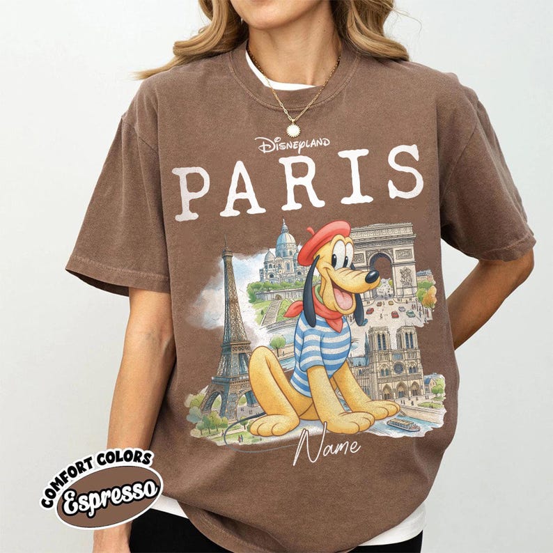 K&ouml;nnte beinhalten: Braunes T-Shirt mit dem Wort "PARIS" in wei&szlig;en Buchstaben und einer Grafik von Pluto vor Pariser Wahrzeichen. Das Shirt hat auch die Worte "Disneyland" &uuml;ber "PARIS" und "Comfort Colors Espresso" darunter.