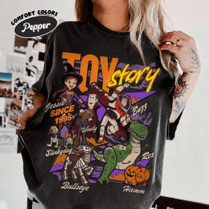 May include: Dark gray Comfort Colors t-shirt featuring a graphic of Toy Story characters, including Jessie, Woody, Buzz, Rex, and Hamm. The design includes the text "TOY STORY" and "Since 1995" in orange and yellow.