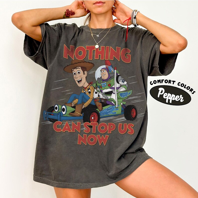 May include: A charcoal gray short-sleeve t-shirt featuring a graphic of Woody and Buzz Lightyear riding a go-kart. The text reads NOTHING CAN STOP US NOW. A circular label on the right side displays the text COMFORT COLORS Pepper.
