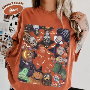 May include: A rust-colored t-shirt featuring a graphic print of various animated characters dressed in Halloween costumes. The design includes characters labeled as Woody, Buzz Lightyear, Jessie, Bo Peep, Rex, Slinky, Forky, Ham, and others.