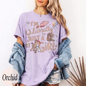 K&ouml;nnte beinhalten: Ein helllila T-Shirt mit dem Text "I'm Literally Just a Girl" und Cowboy-Grafiken. Das Design umfasst ein Hufeisen, einen Cowboyhut, Stiefel und ein Pferd mit einem Cowgirl. Das Shirt hat einen bequemen, l&auml;ssigen Stil.
