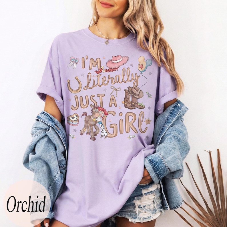 K&ouml;nnte beinhalten: Ein helllila T-Shirt mit dem Text "I'm Literally Just a Girl" und Cowboy-Grafiken. Das Design umfasst ein Hufeisen, einen Cowboyhut, Stiefel und ein Pferd mit einem Cowgirl. Das Shirt hat einen bequemen, l&auml;ssigen Stil.