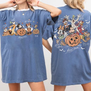 May include: Blue t-shirt featuring a Halloween-themed graphic of Disney characters, including Mickey Mouse, Minnie Mouse, and Goofy, with carved pumpkins. The design is on both the front and back of the shirt, with character signatures.