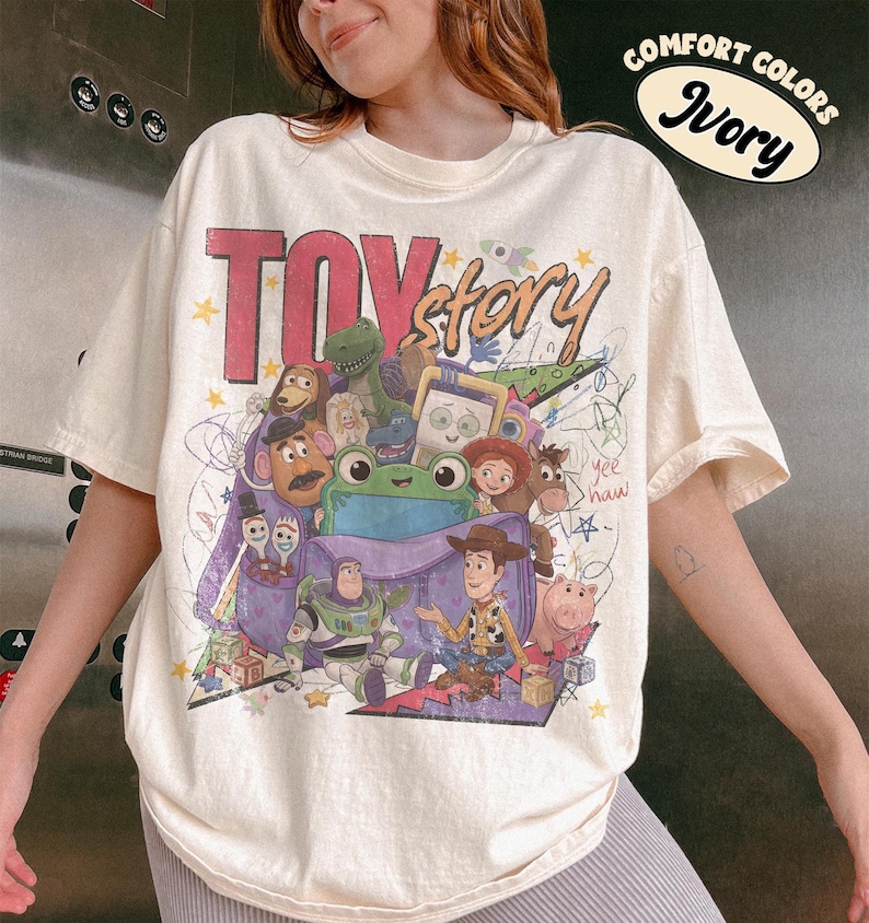 Op de afbeelding: Een ivoorkleurig t-shirt met een grote, kleurrijke grafische print van Toy Story-personages, waaronder Woody, Buzz Lightyear, Rex en Mr. Potato Head. Het ontwerp bevat de tekst "TOY story" in vetgedrukte letters, "yee haw" in cursief en kleurrijke krabbels.