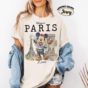 K&ouml;nnte beinhalten: Elfenbeinfarbenes T-Shirt mit dem schwarzen Aufdruck "Disneyland PARIS". Es zeigt eine Cartoon-Mickey-Mouse in Matrosenkleidung, die ein Schokoladeneis h&auml;lt und vor dem Eiffelturm und anderen Pariser Wahrzeichen winkt. Das Shirt ist mit "Comfort Colors" gekennzeichnet.
